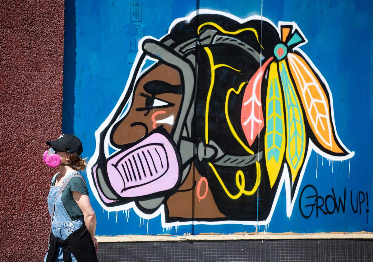 A woman wearing a P100 respirator, amid concerns about COVID-19, walks past artwork of the Chicago Blackhawks NHL hockey team's Native American chief logo wearing a respirator, in the Downtown Eastside of Vancouver, on Friday, April 10, 2020. 