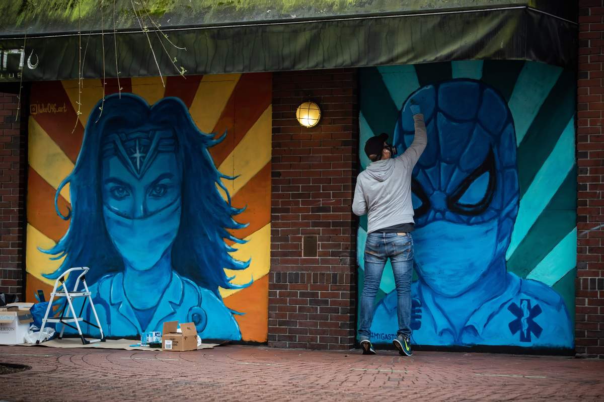Artist Lukas Lundberg touches up a painting of Spiderman depicted as a paramedic while working on a painting of Wonder Woman as a doctor, on the boarded up windows of a closed Gastown business, in Vancouver, on Sunday, April 26, 2020.