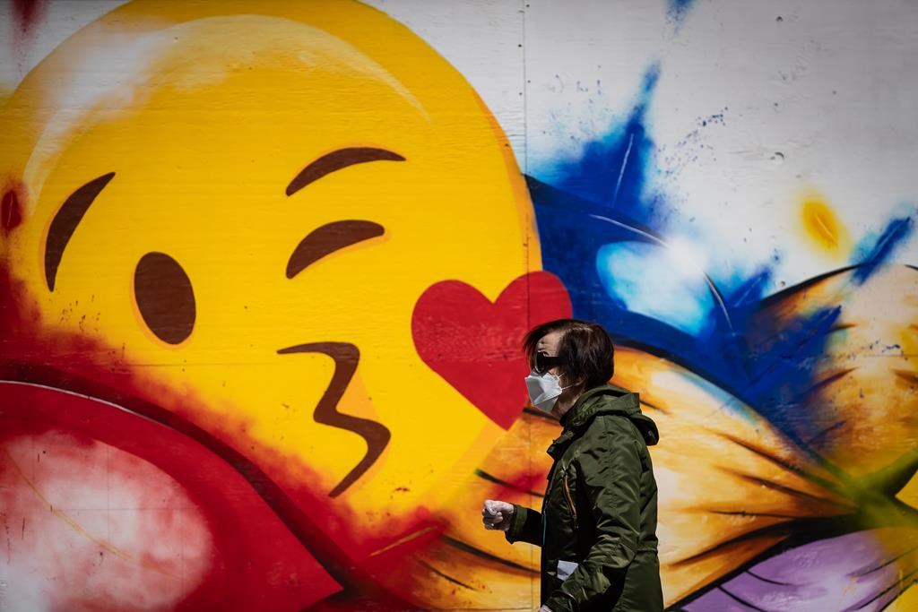 A woman wearing a protective face mask and gloves walks past a large emoji face painted on the boarded up windows of a store on Robson Street, in Vancouver, on Wednesday, May 6, 2020. British Columbia has outlined a gradual reopening of its economy with certain health services, retail outlets, restaurants, salons and museums resuming some operations in mid-May. THE CANADIAN PRESS/Darryl Dyck.