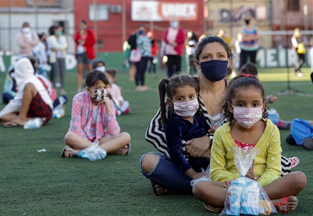 Residents sit on a soccer field as they wait to receive kits equipped with cleaning products and protective face masks for children, provided by a non-governmental organization as a measure to help control the spread of the new coronavirus, in the Paraisopolis slum of Sao Paulo, Brazil, Wednesday, June 24, 2020. With over 100,000 residents, Paraisopolis is one of the areas of Sao Paulo that is most affected by COVID-19. (AP Photo/Andre Penner).