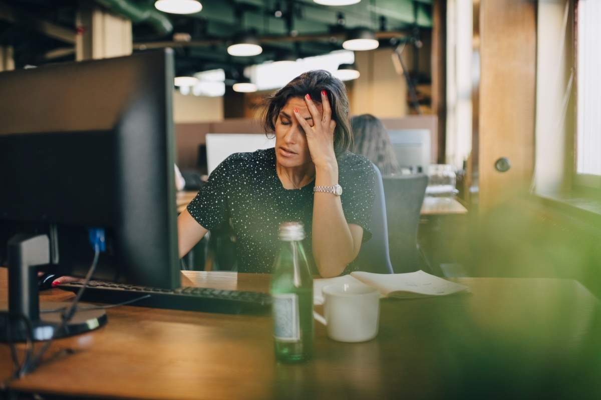 A tired woman sits with head in hand at a computer desk.
