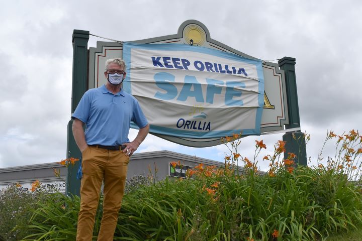 Orillia Mayor Steve Clarke stands in front a city entrance signed, which is covered by a mask promoting safety during COVID-19.