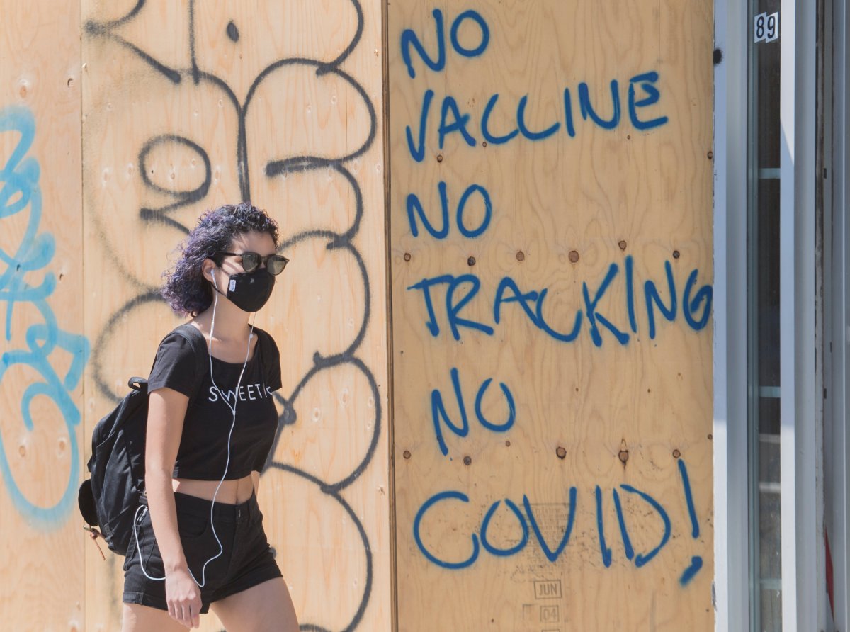 A woman wears a face mask as she passes by graffiti reading 'No vaccine, No tracking, No COVID', in Montreal, Sunday, August 16, 2020, as the COVID-19 pandemic continues in Canada and around the world. 