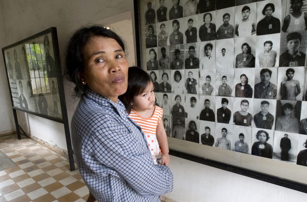 FILE – In this Feb. 12, 2009, file photo, a local woman holds her baby as she stands next to portraits of former prisoners on display at a the notorious former Khmer Rouge prison, S-21, now the Tuol Sleng genocide museum, in Phnom Penh, Cambodia. The Khmer Rouge’s chief jailer, who admitted overseeing the torture and killings of as many as 16,000 Cambodians while running the regime’s most notorious prison, died at a hospital in Cambodia early Wednesday morning, Sept. 2, 2020. Kaing Guek Eav, known as Duch, was 77 and had been serving a life prison term for war crimes and crimes against humanity. (AP Photo/Heng Sinith, File)