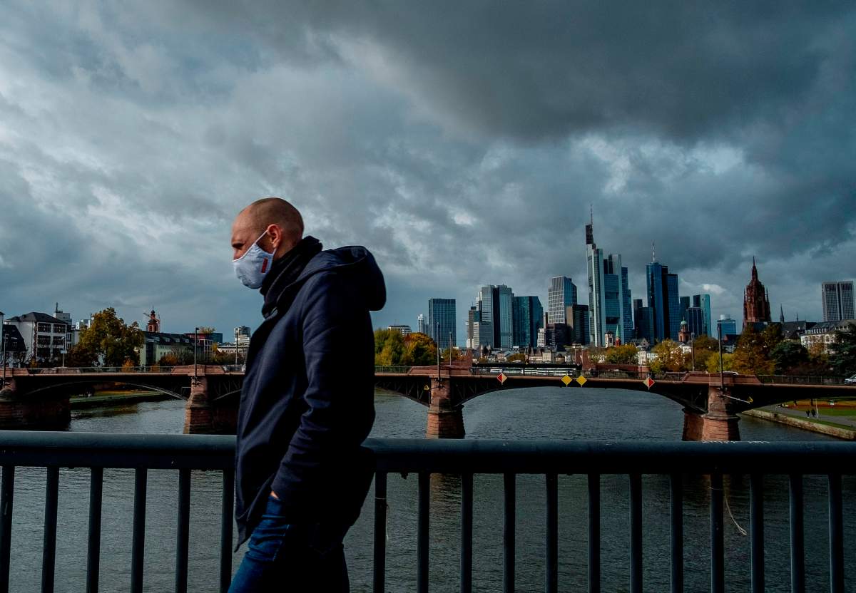 A man wearing a face mask walks over a bridge with the buildings of the banking district in background in Frankfurt, Germany, Thursday, Oct. 29, 2020.