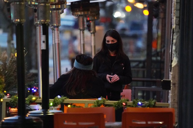A waitperson wears a face mask while tending to a patron sitting in the outdoor patio of a sushi restaurant, late Monday, Dec. 28, 2020, in downtown Denver. 
