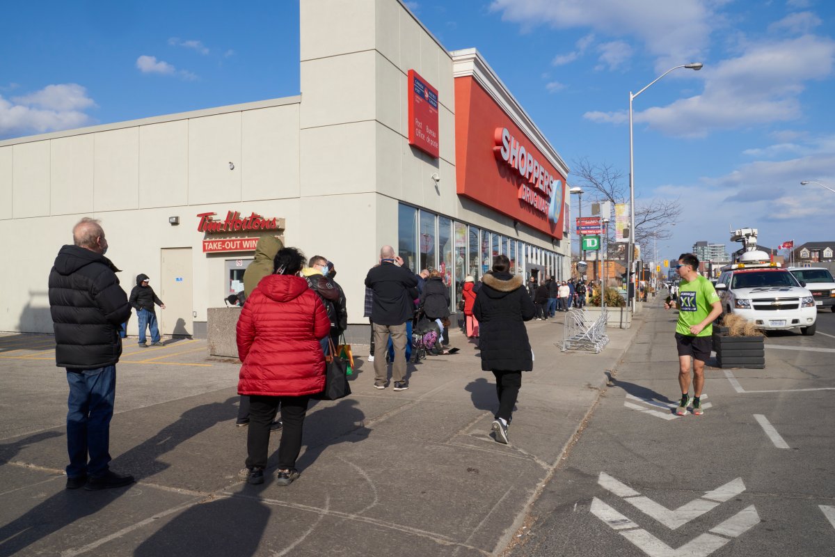 A man runs past people in the age group of 60-64 as they line up to get their Oxford-AstraZeneca COVID-19 vaccinations at the Shoppers Drug Mart at Danforth and Coxwell, in Toronto on March 11, 2021, exactly one year to the day that the World Health Organization declared a global pandemic. 