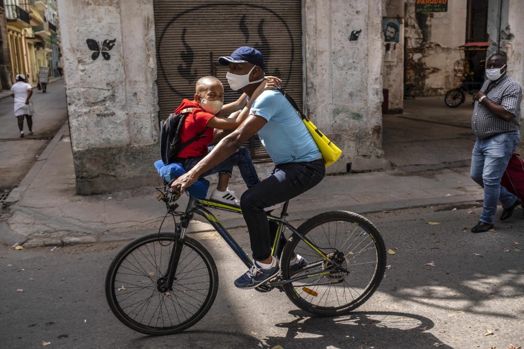 A man rides a bicycle with a child on his handlebars in Havana, Cuba, Friday, March 19, 2021, amid the COVID-19 pandemic. (AP Photo/Ramon Espinosa).