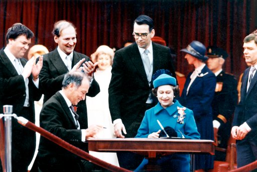 Prime Minister Trudeau and the Queen prepare to sign the Constitution proclamation as Consumer Affairs Minister Ouellet (left), Secretary of State Regan and Privy Council Clerk Pitfield look on.