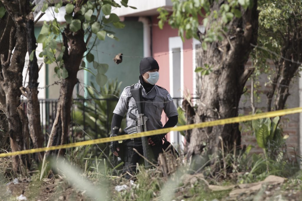 A police officer stands guard behind police tape, near the house of a former policeman where forensic teams work to exhume several graves in Chalchuapa, El Salvador, Wednesday, May 19, 2021. The search at the home of the former policeman arrested last week for the murder of two women has turned up 10 other bodies, prosecutors said Friday, revealing the existence of a murder ring that allegedly killed as many as 13 people. (AP Photo/Salvador Melendez).
