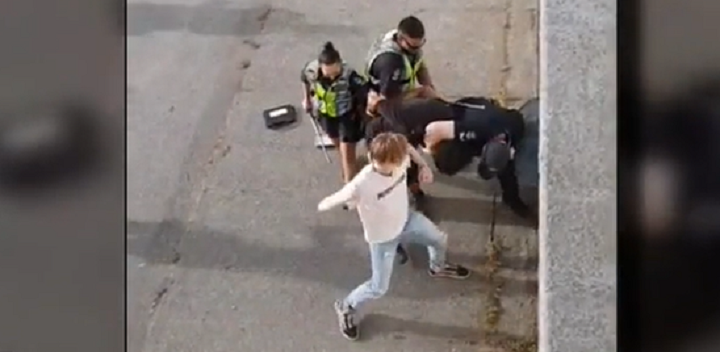A member of a crowd approaches police with his fist raised as officers take a handcuffed man to the ground. 