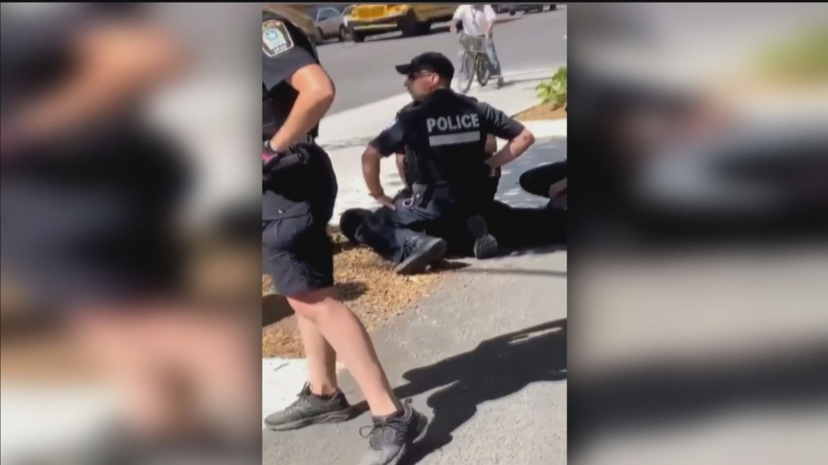 The video shows two police kneeling over a young Black male who is lying on the ground in front of a bus stop, with one officer placing his knees on the person's neck and shoulders.