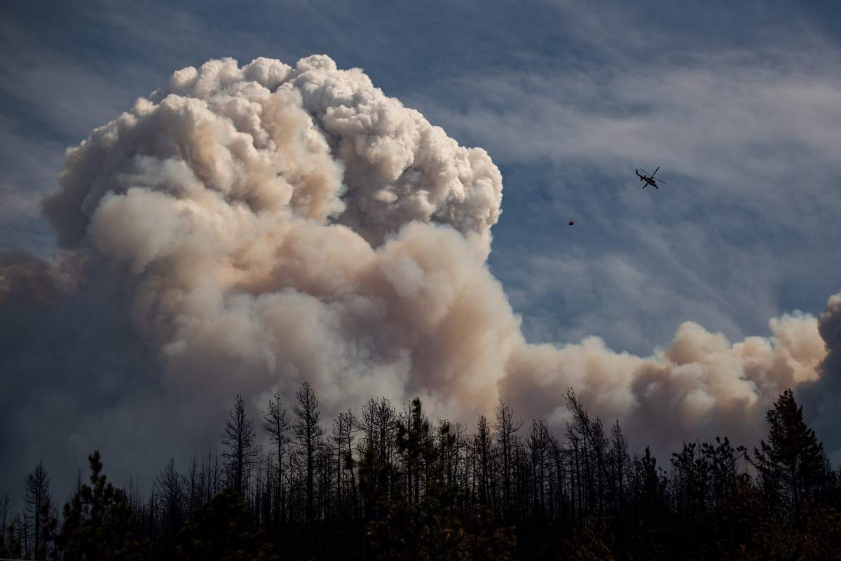 A helicopter carrying a water bucket flies past a pyrocumulus cloud, also known as a fire cloud, produced by the Lytton Creek wildfire burning in the mountains above Lytton, B.C., on Sunday, August 15, 2021. 