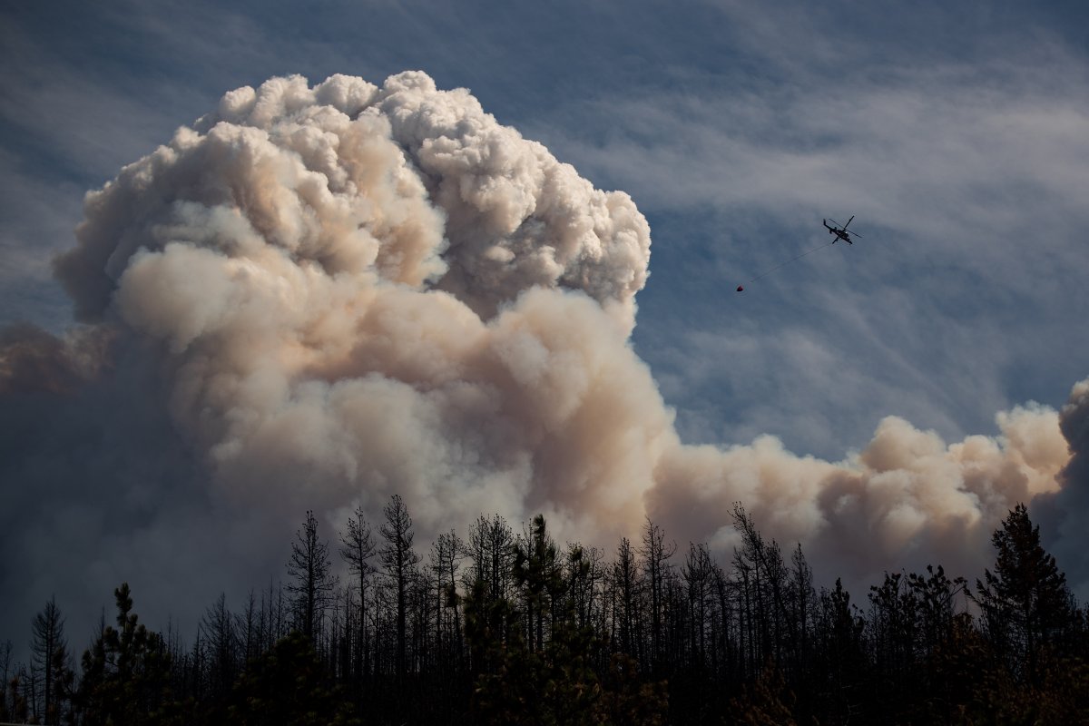 A helicopter carrying a water bucket flies past a pyrocumulus cloud, also known as a fire cloud, produced by the Lytton Creek wildfire burning in the mountains above Lytton, B.C., on Sunday, August 15, 2021. 