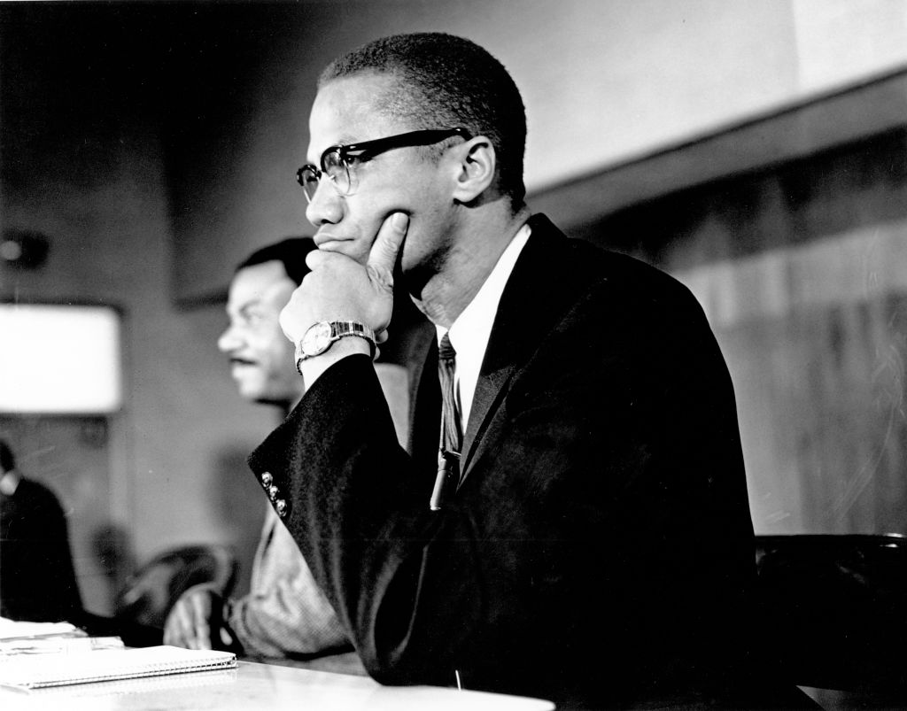 A photo of Malcolm X sitting at a table with his chin resting in his hands staring away from the camera