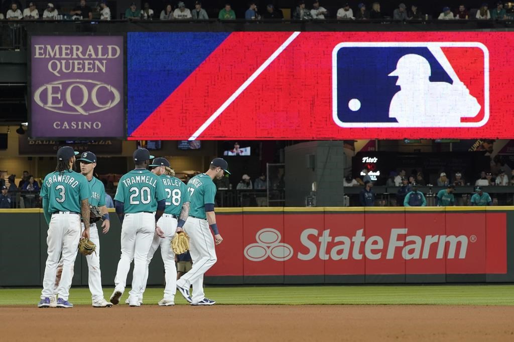 FILE - Seattle Mariners gather as the MLB logo is shown during a review of an attempted catch by right fielder Mitch Haniger of a ball hit by Tampa Bay Rays' Ji-Man Choi that was originally called an out during the ninth inning of a baseball game Friday, June 18, 2021, in Seattle. The call was overturned. The Mariners won 5-1. The clock ticked down toward the expiration of Major League Baseball’s collective bargaining agreement at 11:59 p.m. EST Wednesday night, Dec. 1, 2021, and what was likely to be a management lockout ending the sport’s labor peace at over 26 1/2 years. (AP Photo/Ted S. Warren).