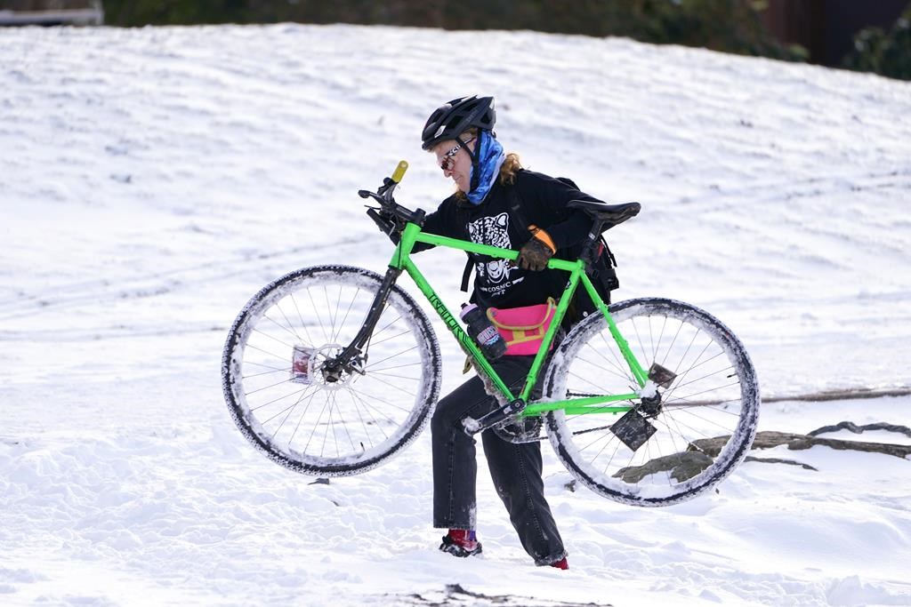 Apollo Leonard hefts his bike through snow too deep to ride in after stopping while out for a winter ride, Tuesday, Dec. 28, 2021, in Bellingham, Wash. Snow, ice and unseasonable cold in the Pacific Northwest and the Sierra Nevada are continuing to disrupt traffic, cause closures and force people to find refuge in emergency warming shelters.