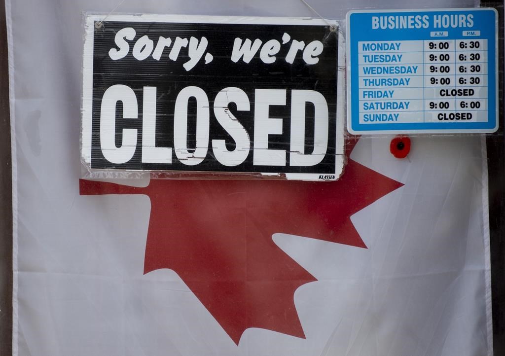 A "Sorry, we're closed" sign in front of a hanging Canadian flag in a store window, next to a blue and white sticker showing business hours.