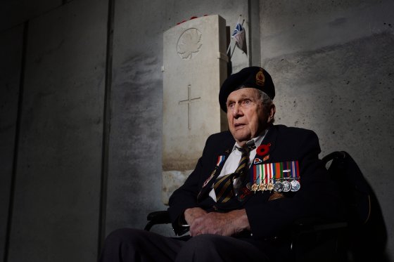 Second World War veteran Fred Arsenault sits in a wheelchair before a grey headstone, staring off into the distance. He wears a military uniform with colourful badges pinned to his chest above his heart, and a small cap upon his head. His face is lined with age, and solemn.