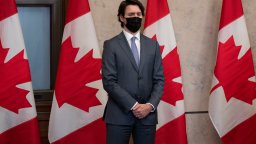 Prime Minister Justin Trudeau stands in front of a row of red and white Canadian flags. He stares off ahead, wearing a black face mask and a dark suit, with his hands clasped in front of him.
