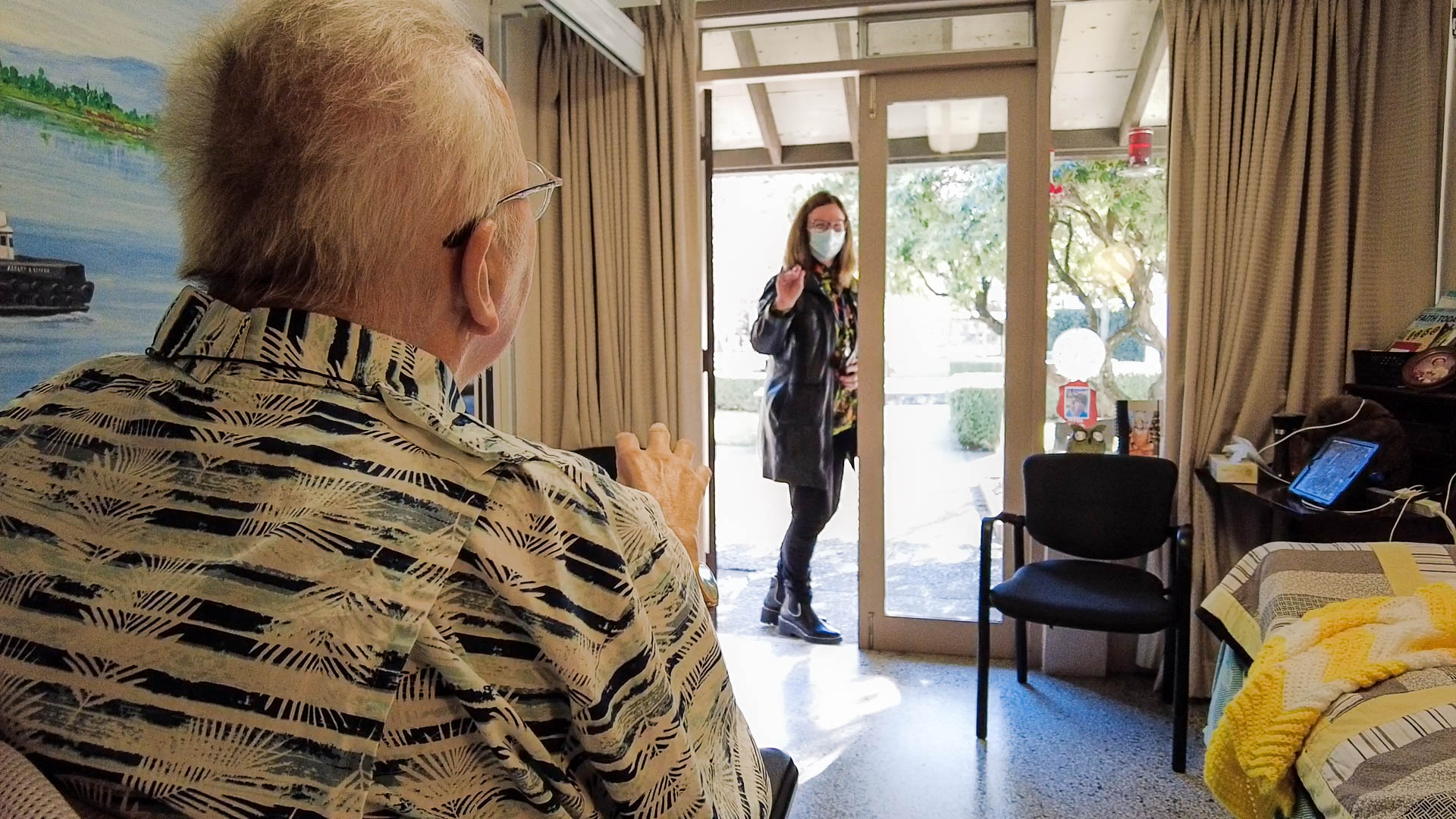 A white woman with shoulder-length hair and glasses waves goodbye to a man seated with his back to the camera.