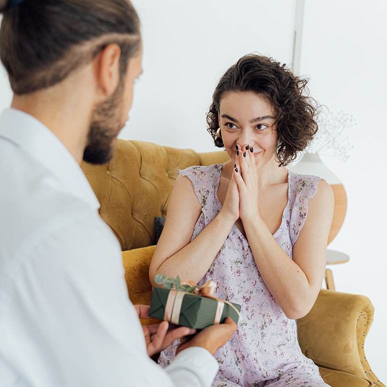 Couple seated, with one partner gifting a wrapped box to another excited partner