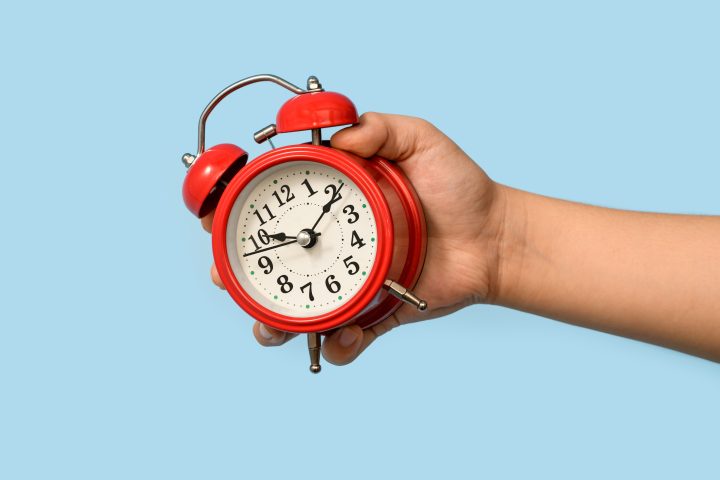 Cropped Hand Of Boy Holding Alarm Clock On Blue Background.
