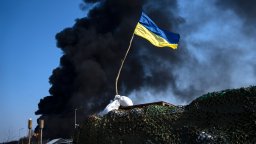 A blue and yellow Ukrainian flag flies alone against a plume of black, billowing smoke caused by a Russian attack.
