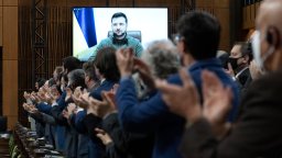 Ukrainian President Volodymyr Zelenskyy is visible on a screen behind a long row of standing Canadian MPs applauding him.