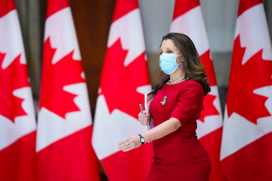 Finance Minister Chrystia Freeland is mid-stride in front of a wall of Canadian flags, wearing a red dress and a blue face mask.