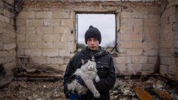 A young Ukrainian boy looks straight into the camera while holding his pet cat in the ruins of his family home, destroyed by Russian forces.