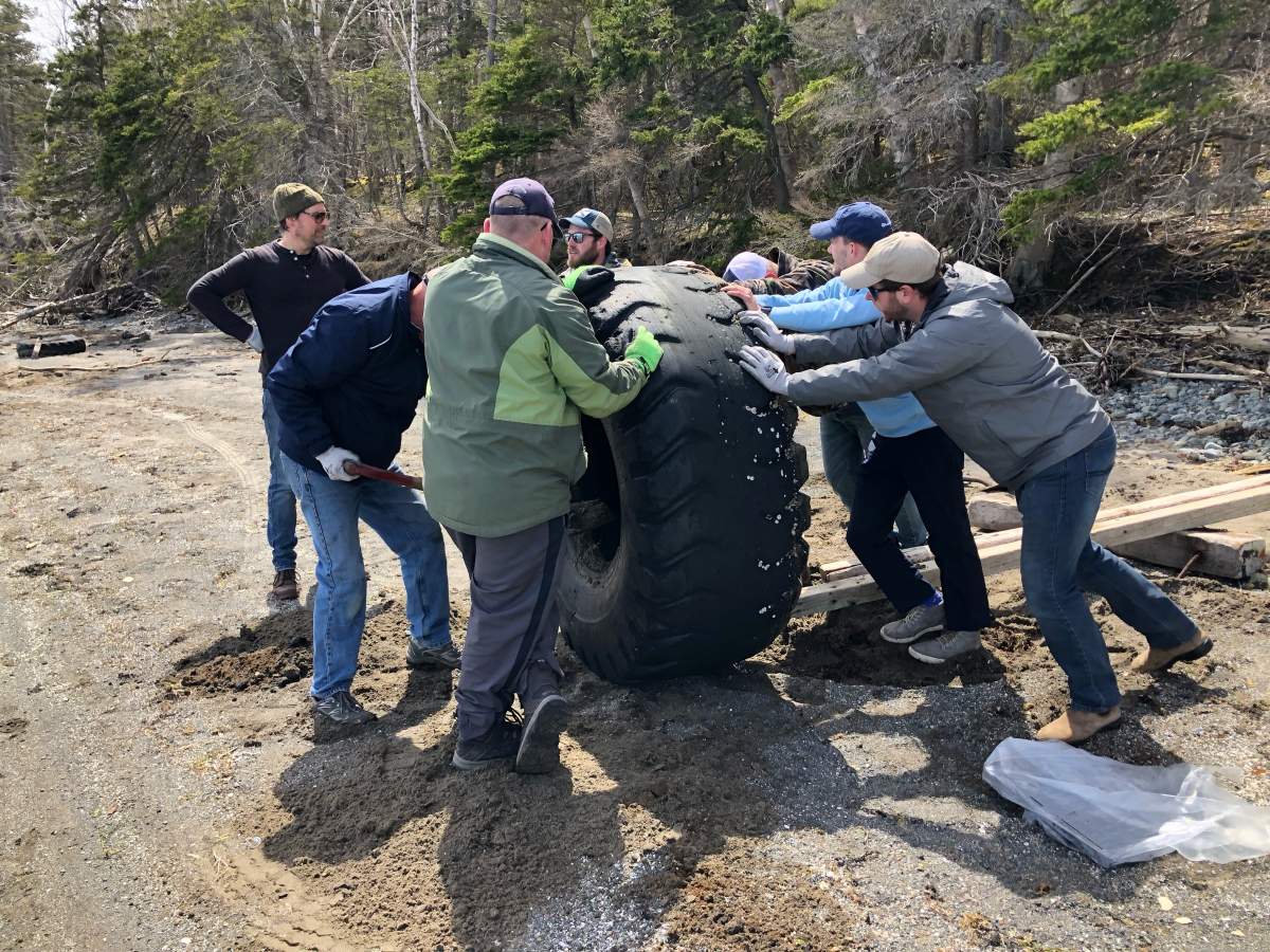 Volunteers are pushing a large tire across a beach in Nova Scotia