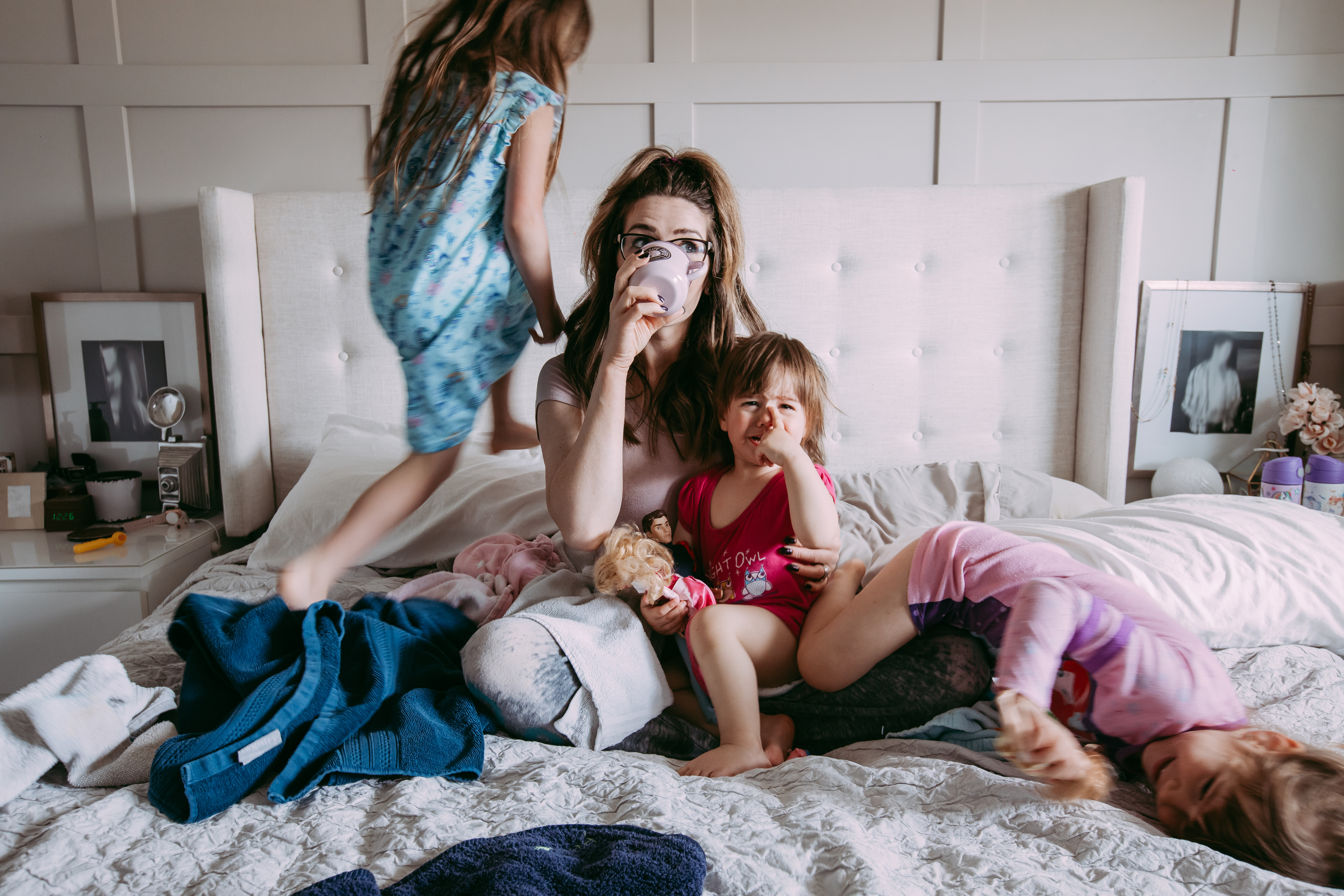 A mom sips coffee in bed while her children climb on her.