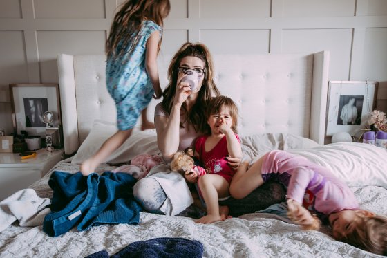 A mom sips coffee in bed while her children climb on her.