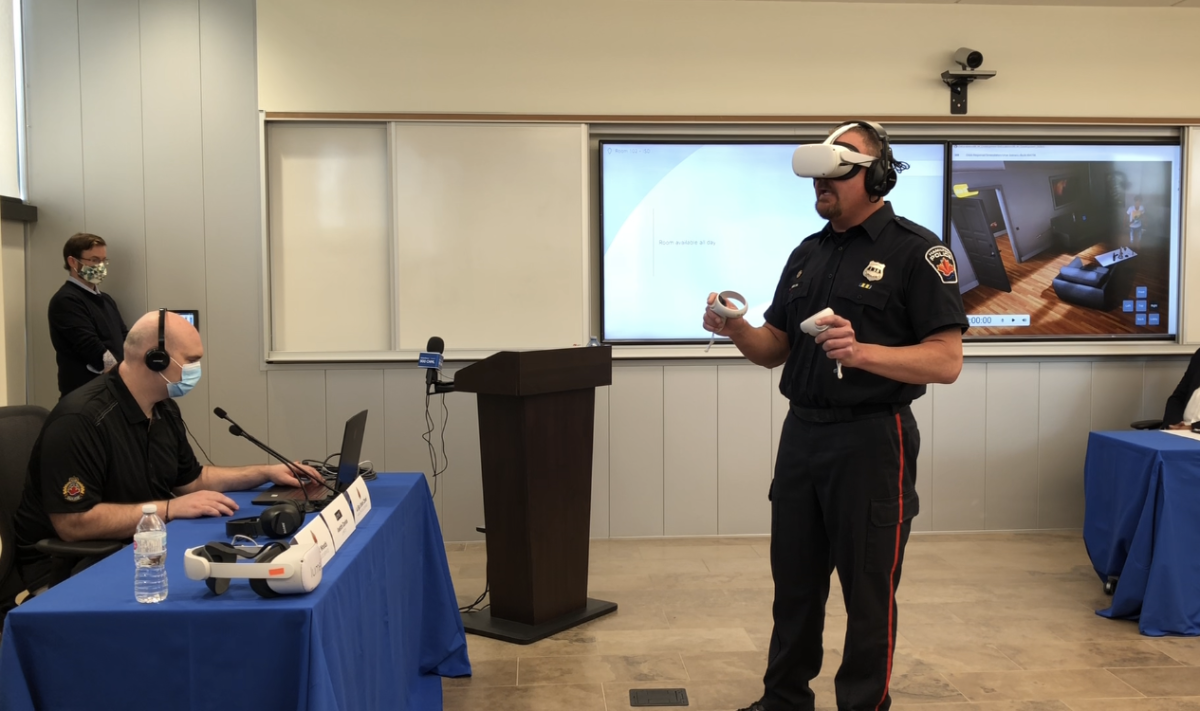 A Hamilton police officer wearing virtual reality technology stands in a room with a screen showing what he sees behind him. There is a man to the left on a laptop controlling the simulation and another man standing behind him.