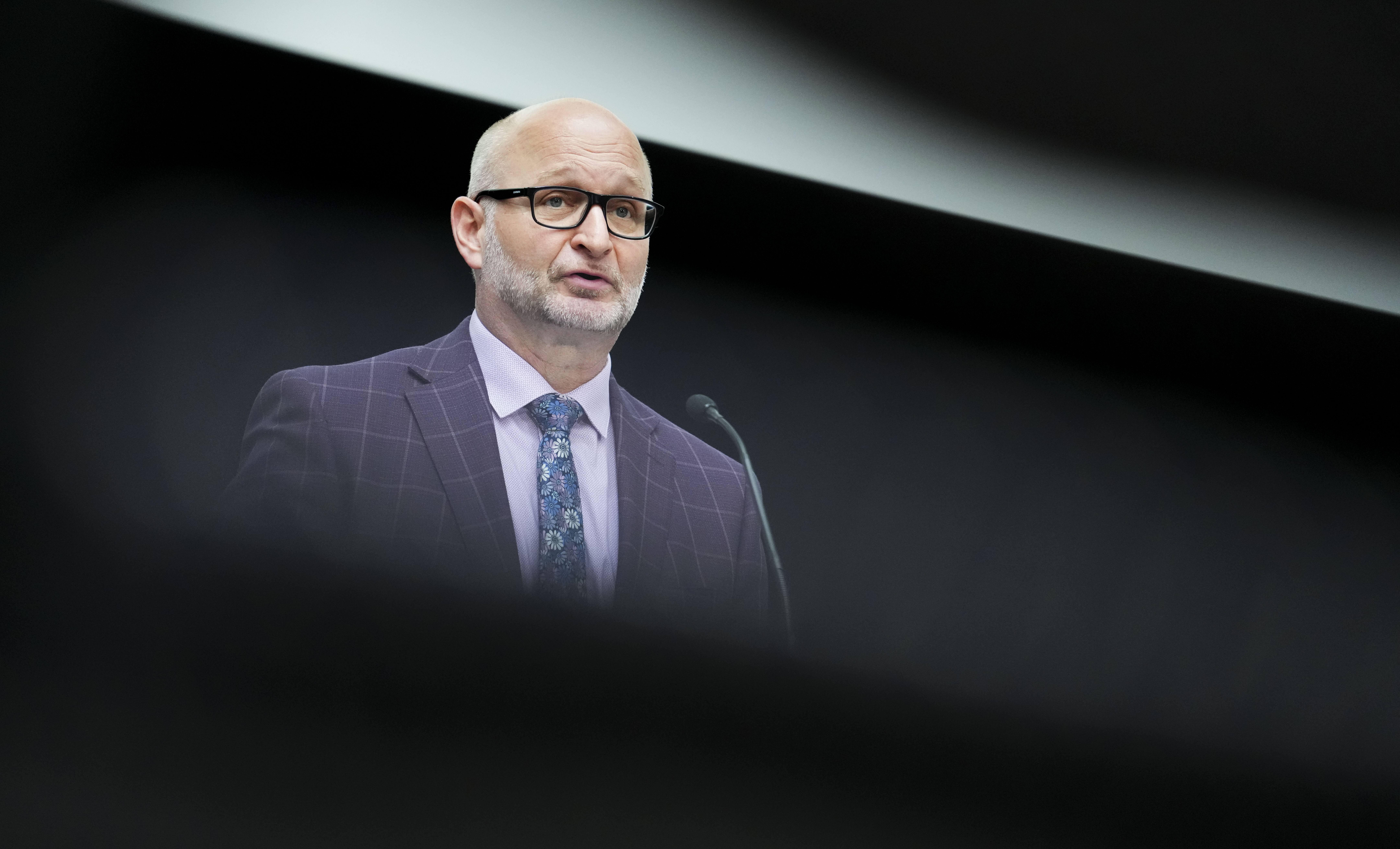 Justice Minister David Lametti stands at a press conference. He wears a grey suit against a black background.