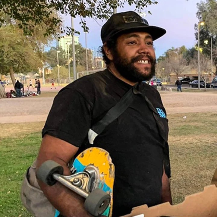 Sean Bickings is seen holding a skateboard in what appears to be a park, with grass and trees in the background.
