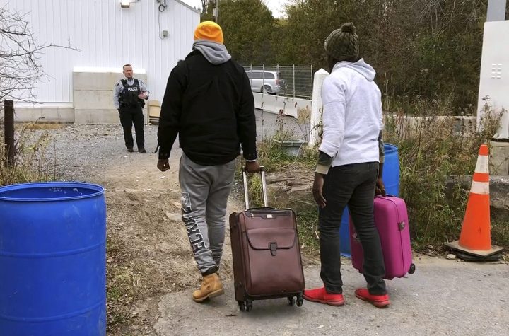 In this Nov. 4, 2019, photo, two men from Nigeria cross the U.S. border at Roxham Road in Champlain, N.Y., into Canada where Royal Canadian Mounted Police stand, rear, in Saint-Bernard-de-Lacolle, Quebec. (AP Photo/Wilson Ring).
