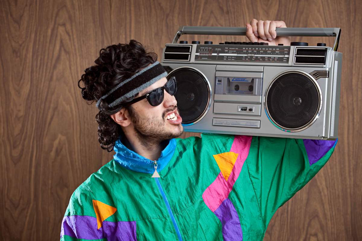 A man in an 80s costume and mullet wig holds a boom box on his shoulder.