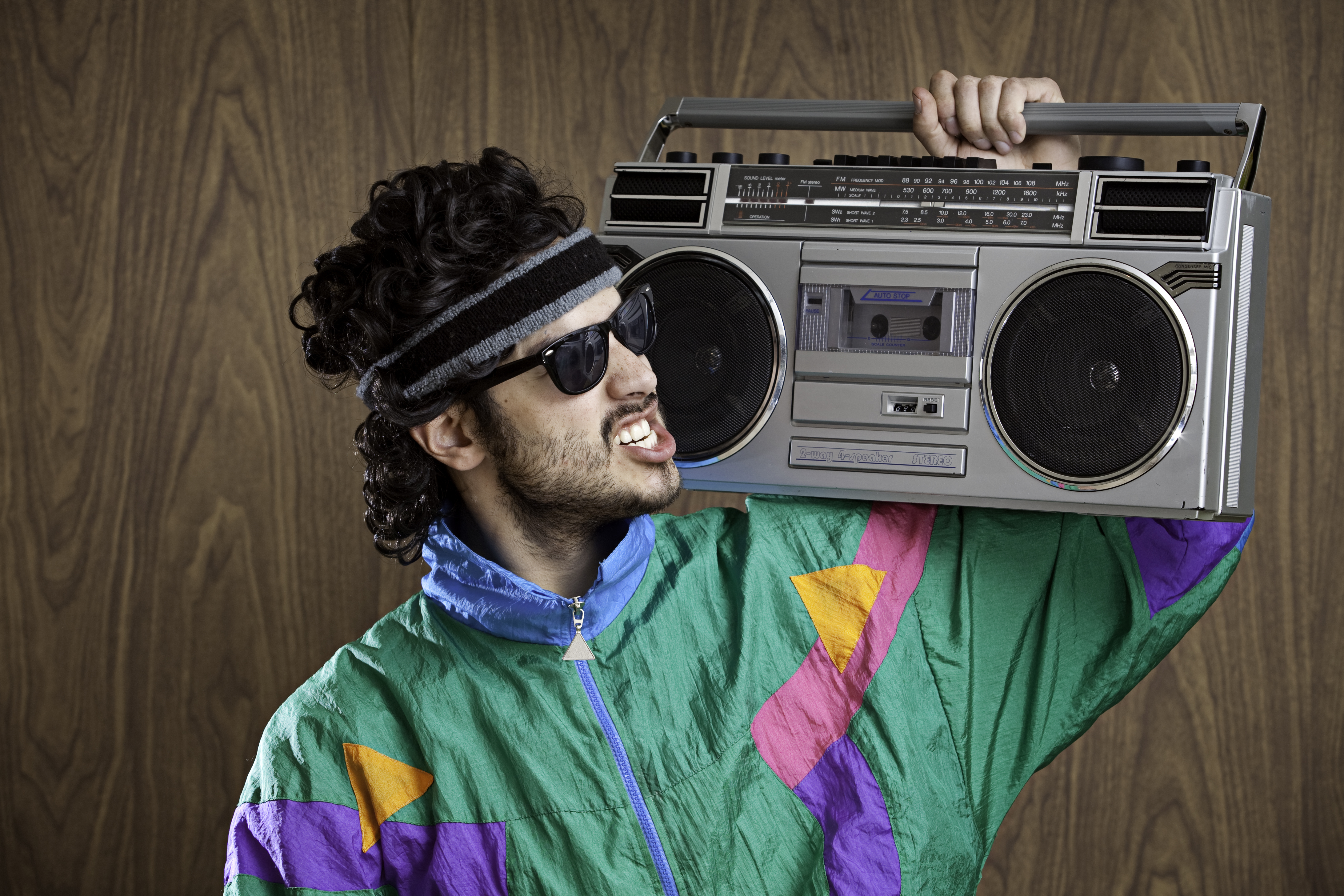 A man in an 80s costume and mullet wig holds a boom box on his shoulder.