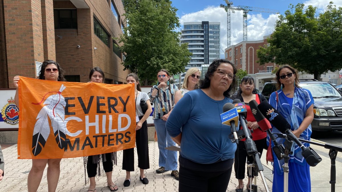 Members of the Indigenous community gather in front of the Hamilton police station, speaking to media and holding an orange 'EVERY CHILD MATTERS' banner.