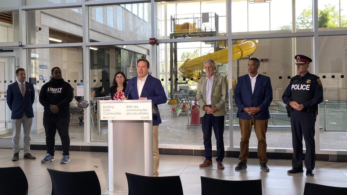 Councillor John Paul Danko, Liberty For Youth's Abubakar Kayiranga, Procurement Minister Filomena Tassi, Mayor Fred Eisenberger, MP Matthew Green, and Chief Frank Bergen stand behind Public Safety Minister Marco Mendocino during an announcement at Westmount Recreation Centre. They're all in front of a glass window that looks out onto an indoor pool with a waterslide.