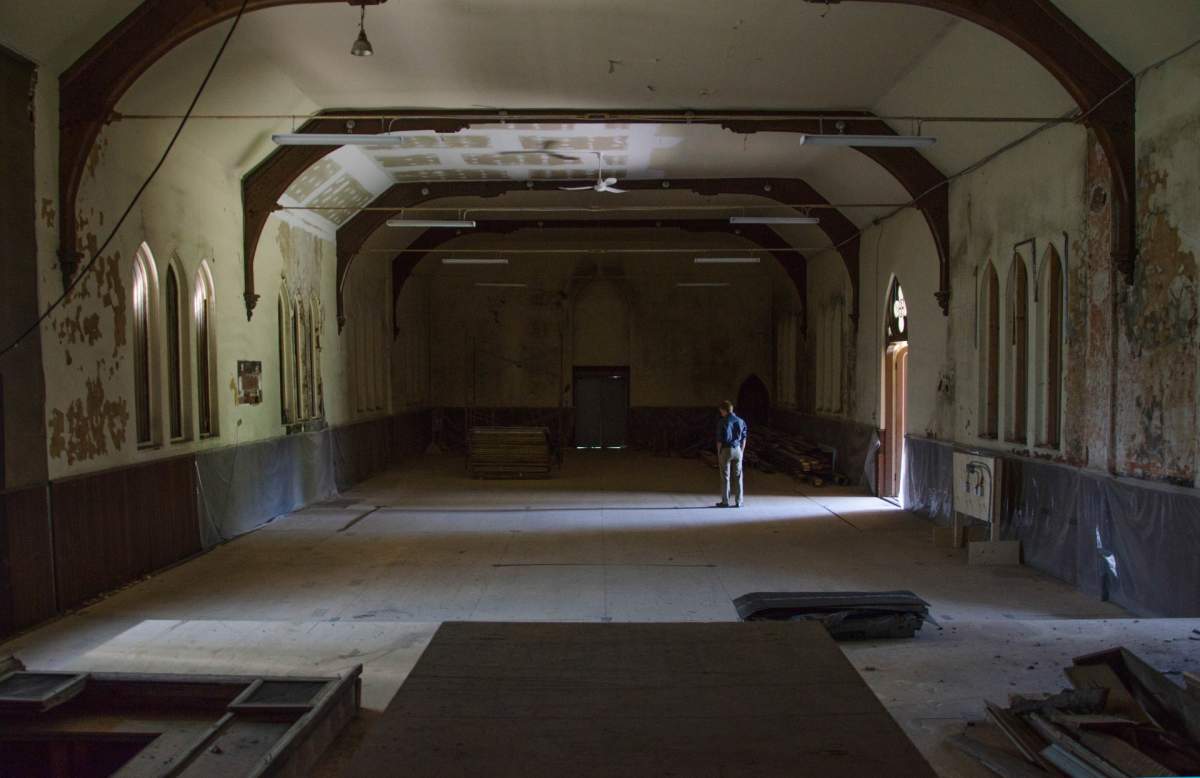 A man stands inside a large dark and empty room, the inside of an old church, with sunlight streaming in from an open door.