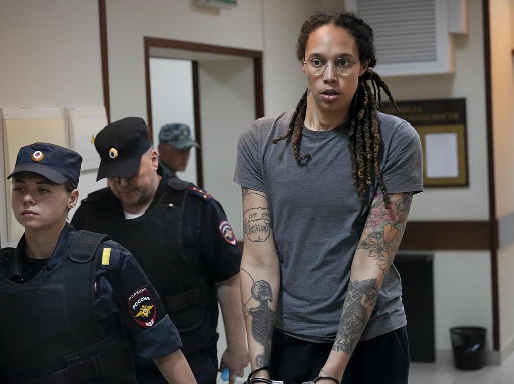 Brittney Griner in a grey tee and handcuffs, escorted by considerably shorter uniformed officers.