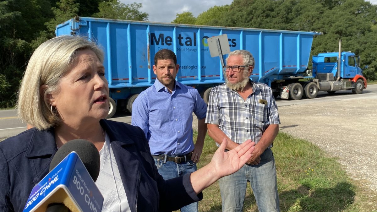 Andrea Horwath stands in front of residents at Highway 52 and Powerline Road West, while a large blue freight truck rolls by in the background.
