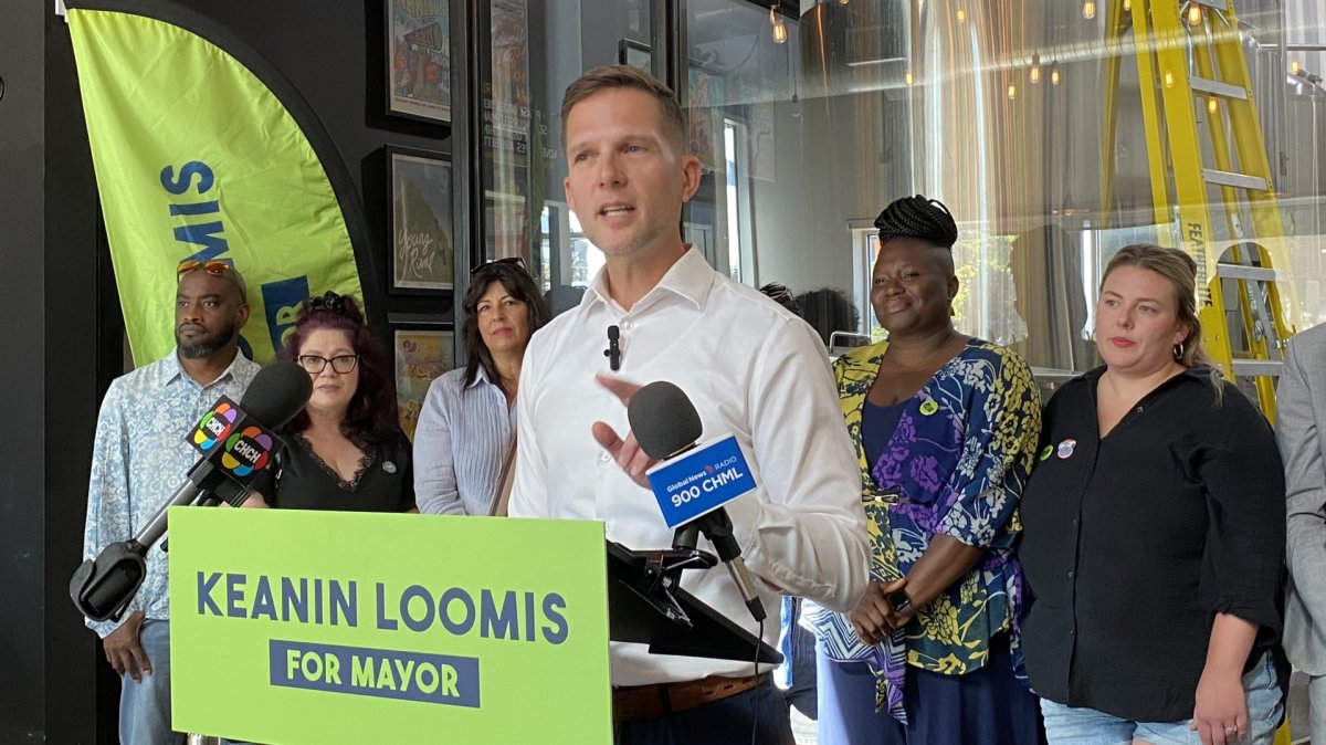 Keanin Loomis stands at a podium, while local business owners and entrepreneurs stand behind him during a media availability at Merit Brewing on James Street North.