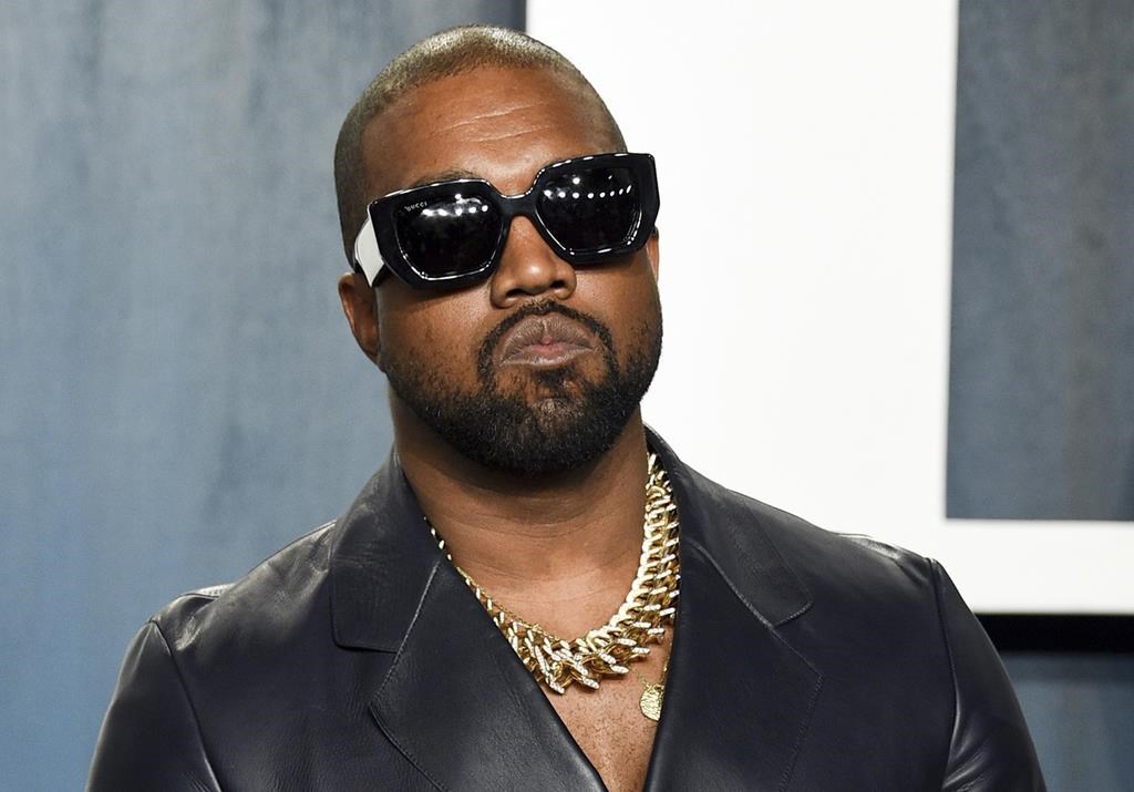 Kanye West on a red carpet. He is wearing dark sunglasses, a gold chain and a suit blazer.