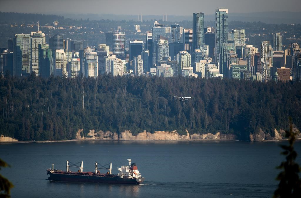 A bulk carrier cargo ship travels into port as a Harbour Air seaplane flies towards Stanley Park and the downtown skyline, in Vancouver, on Wednesday, July 27, 2022.