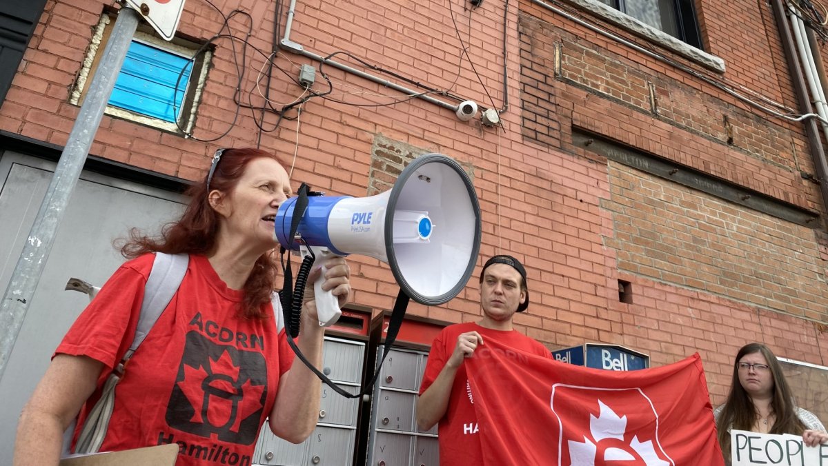 A woman wearing a red shirt holds a megaphone and speaks passionately into it. Standing beside her is a man wearing a red shirt and holding up a red flag, next to another woman who is holding a sign. All three are standing in front of a brick wall outside of an apartment building.