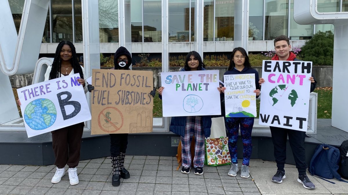 Five students from a Hamilton high school stand in front of the Hamilton sign at city hall, holding signs with messages about the climate crisis.